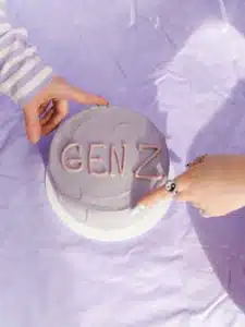 High angle view of a purple cake with 'Gen Z' icing touched by hands on a lavender surface.