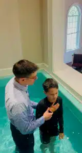 Man performing a baptism on a young boy in a church, with an arched window and pews visible in the background.