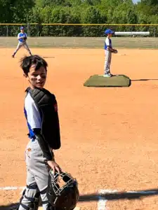 Young baseball catcher in chest protector and helmet on an orange dirt field, with teammates in the background near the pitcher's mound.