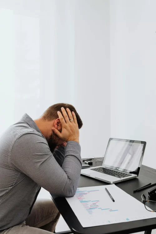 A frustrated man sits at a desk with a laptop and charts, showing work stress.