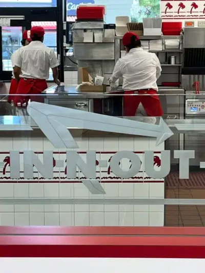 Inside an In-N-Out Burger, two employees in white shirts and red aprons work behind a stainless steel counter with kitchen equipment; the In-N-Out logo appears on the glass in front.