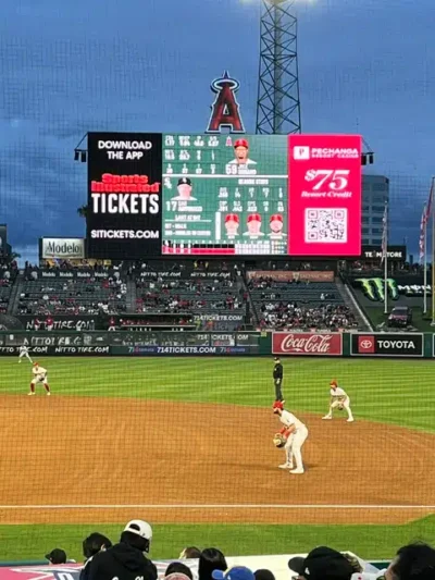 Baseball game at a stadium with a large electronic scoreboard displaying lineups and ads; Angels logo atop the board; batter at the plate.