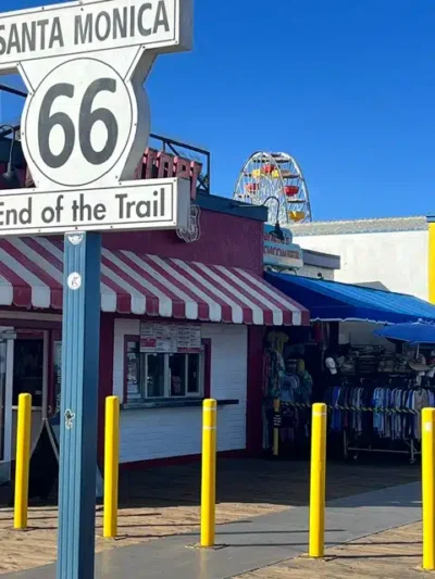 Route 66 sign saying 'Santa Monica' and 'End of the Trail' in front of a striped awning shop and a distant Ferris wheel in clear blue sky.