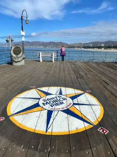 Compass rose painted on the wooden pier deck reading Santa Monica Pier, with a person looking out at the ocean and distant hills.