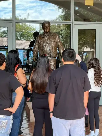 Bronze statue of a man in a jacket standing inside a glass-walled entrance, as a group of people watches from behind.