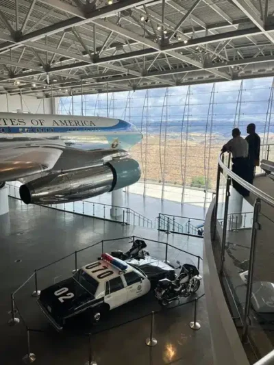 Museum hangar displaying a large aircraft nose and engines, a black-and-white police car, and motorcycles on the floor, with visitors on a curved mezzanine above.