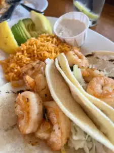 Plate of shrimp tacos with cabbage, cilantro, and soft tortillas, plus citrusy pineapple, avocado slices, and seasoned rice in the background.