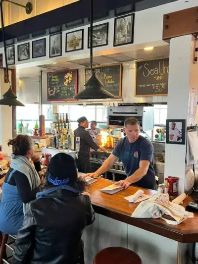 Casual seafood cafe scene with a bar counter where a server in a blue shirt takes a order from customers seated on stools; kitchen activity visible behind, chalkboard menus and framed photos above.