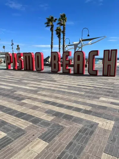 Red 3D letters spell 'Pismo Beach' on a striped brick promenade with palm trees and the ocean in the background.