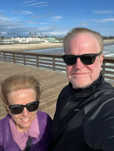 Two people wearing sunglasses take a sunny selfie on a wooden pier, with a beach, ocean, and palm trees in the background.