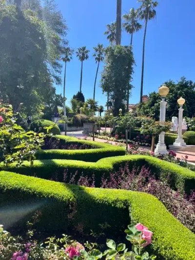 Formal garden with neatly trimmed hedges, blooming flowers, and tall palm trees under a blue sky.