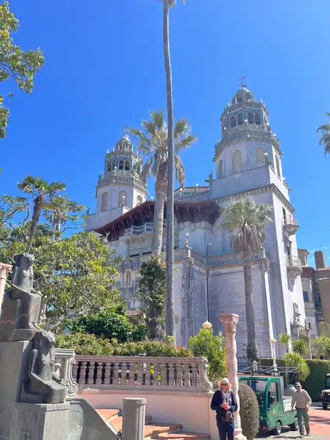 Historic white church with twin domes and ornate architecture, surrounded by palm trees against a bright blue sky, with a garden foreground and a small vehicle nearby.