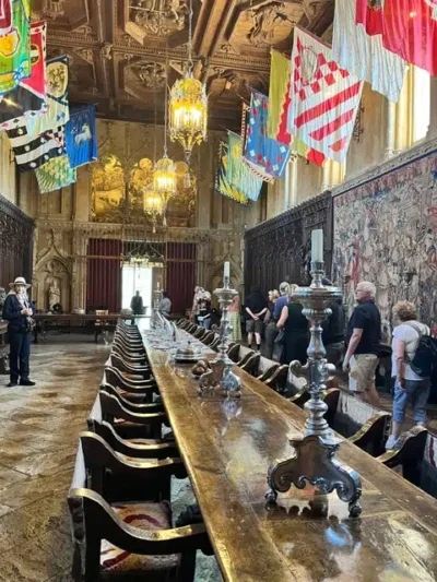 Dining Room at Hearst Castle - TruFlex Media Long wooden banquet table in a grand hall with hanging colorful flags, chandeliers, and visitors touring the room, left a guide with hat stands nearby.
