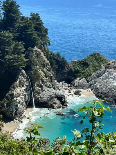 Turquoise cove with rocky cliffs, a small waterfall, and evergreen trees overlooking the blue ocean.