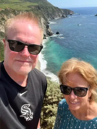 A smiling couple wearing sunglasses takes a selfie on a cliff overlooking turquoise sea and rocky coastline.