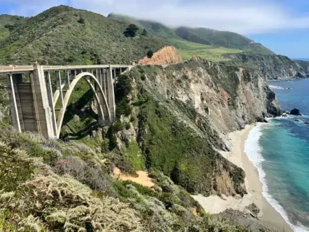 Concrete arch bridge spans a steep coastal ravine, with green hills, rugged cliffs, and a small sandy beach below the turquoise sea.