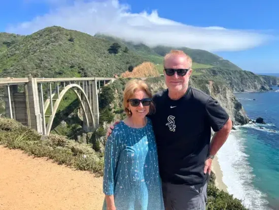 Couple wearing sunglasses poses on a cliff, with a curved coastal bridge and blue ocean behind them under a clear sky.