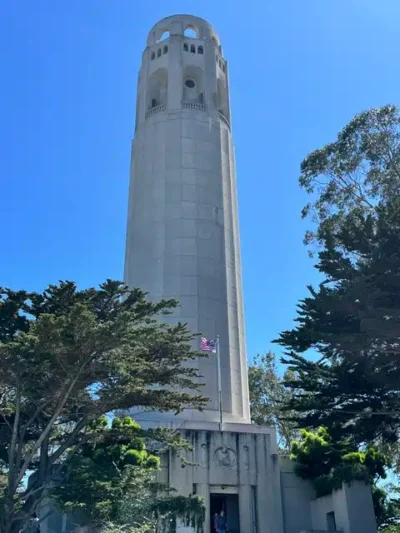 Tall white tower with arched openings at the top, surrounded by trees under a blue sky; American flag at its base.