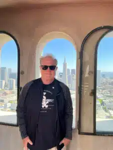 IMG_7348 - TruFlex Media Man wearing sunglasses and a black jacket stands in front of arched windows with a city skyline behind him.