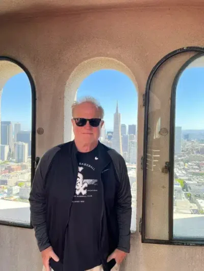 Man wearing a black jacket and sunglasses poses inside a stone arched-window tower with a city skyline and blue sky behind him.