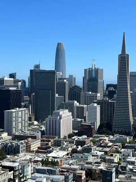 City skyline with tall modern skyscrapers against a clear blue sky; a rounded tower and a sharp spire stand among densely packed buildings in the foreground.