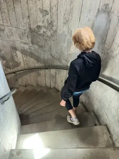 Person with blonde hair, wearing a black hoodie and shorts, descends a concrete spiral staircase with curved metal handrails along a rough concrete wall.