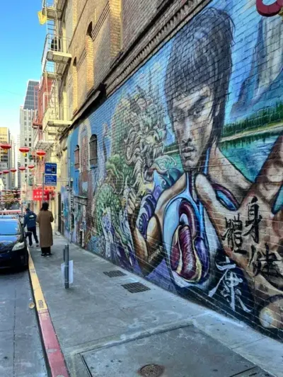 Narrow city street with a tall brick wall covered in a colorful mural featuring a man's portrait and a dragon-like design; pedestrians and parked cars along the sidewalk.