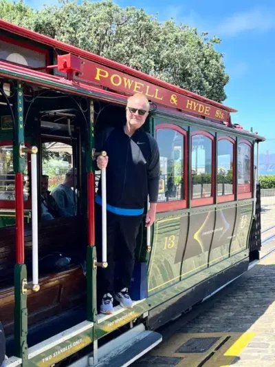 Man in black outfit and sunglasses stands in the open doorway of a vintage Powell & Hyde Street trolley, holding a pole on a sunny day.