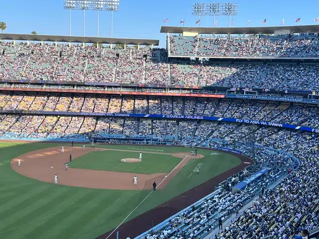 Baseball game in progress at a large stadium with players on the field and a crowded audience under a blue sky.