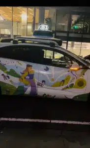 White van with a colorful mural featuring people and abstract shapes, parked at night in a lot with a modern building in the background; roof-mounted light bar visible on top of the vehicle.