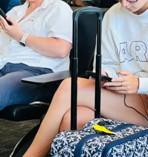 Focused - TruFlex Media Two travelers seated in an airport lounge with a rolling suitcase between them; both look at their phones.