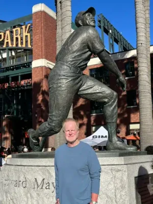 Willie Mays Statue - TruFlex Media Man in a blue long-sleeve shirt posing in front of a large bronze baseball pitcher statue outdoors, with palm trees and a brick building in the background and'PARK' signage nearby.