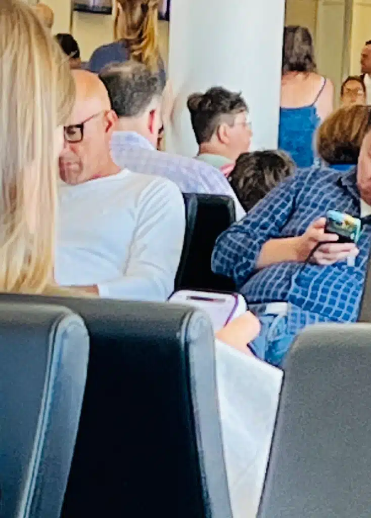 Locked In - TruFlex Media People seated in a waiting area; a man in a white shirt looks down while others nearby, including a man in a blue plaid shirt checking his phone.