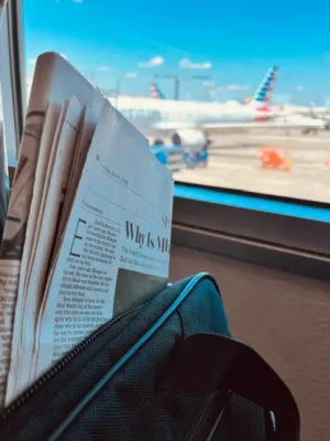 Old School - TruFlex Media Newspaper sticking out of a teal bag at an airport gate, with airplanes on the tarmac outside the window.