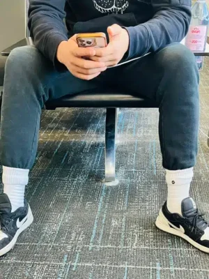 Phone in Hand Always - TruFlex Media Person seated on a chair, wearing dark joggers and sneakers, and holding a smartphone with both hands.