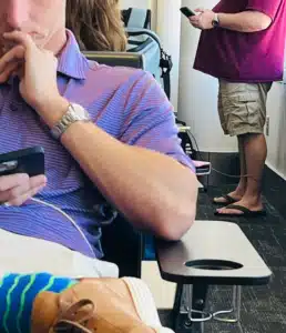 Person in a purple striped polo sits and looks at a smartphone in a waiting area as others stand in the background with phones nearby.
