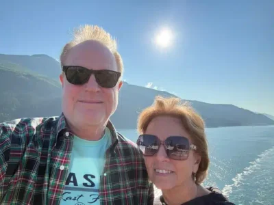 Big Sun - TruFlex Media Smiling man and woman wearing sunglasses aboard a boat, with a blue lake, mountains, and a bright sun in the background.