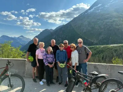 Bike Buddies in Mountains - TruFlex Media Eight adults pose for a group photo on a mountain overlook with two bicycles in the foreground; blue sky, clouds, and snow-capped peaks create a scenic backdrop as they smile at the camera.