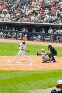Mune Homers - TruFlex Media Baseball game action: batter in white swings at a pitch as catcher and umpire crouch behind home plate with a crowded stands backdrop.